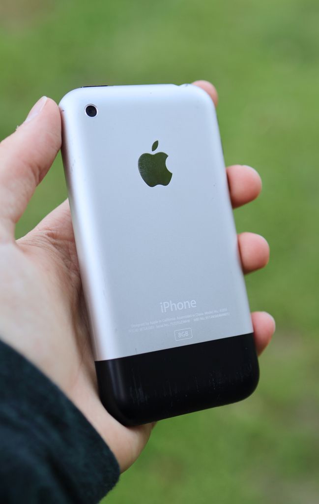 Close-up of a person holding a classic Apple iPhone outdoors on a blurred background.